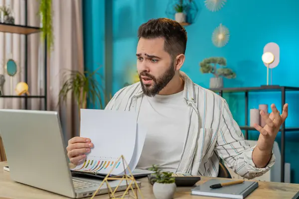 Frustrated upset businessman works at office desk laptop calculate looking at bills paperwork reports documents gets angry frustrated stress bankruptcy financial problem. Freelancer at home. Lifestyle
