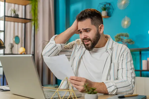 Amazed freelancer man opening envelope reading bad news in letter feels upset. Financial problems, bank debt bill for pay, tax invoice or bankruptcy notification. Guy at home office table. Lifestyle