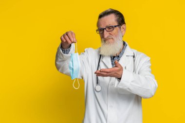 Yaşlı doktor kardiyolog maskesini çıkartıyor. Grip tedavisi. Salgın karantina sona eriyor. Eczacı eczanesi. Yaşlı büyükbaba sarı arka planda izole edilmiş.