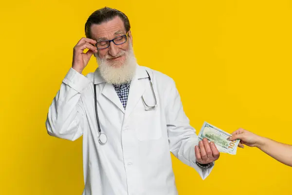Senior speaker doctor cardiologist man receiving bribe from patient taking money from client hiding cash into pocket. Illegal corruption in medicine. Mature apothecary grandfather on yellow background