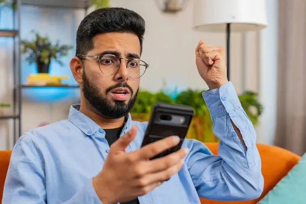 Shocked sad Indian young man using smartphone reading negative message feels annoyed sitting on sofa in living room. Arabian guy with hand on head having gadget trouble at home. App crash concept