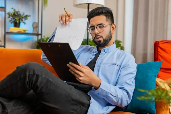 Happy Indian man freelancer work with documents on home couch reading information business papers prepare financial report. Young Hispanic guy accountant analyzing graph documents, calculate taxes.