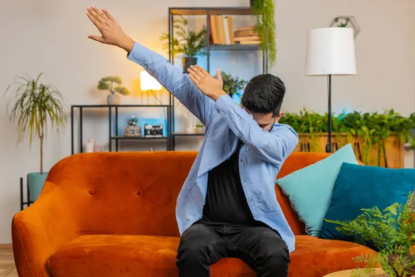 stock image Trendy cheerful positive Indian Arabian man having fun dancing, dabbing raising hands making dub winner dance gesture celebrating victory at home. Happy bearded Indian guy in living room on sofa.