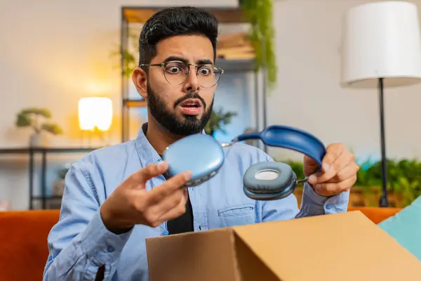 Dissatisfied annoyed Indian male customer open cardboard box receive damaged broken wireless headphones, shocked young man consumer having problem complaint frustrated with bad order delivery at home.