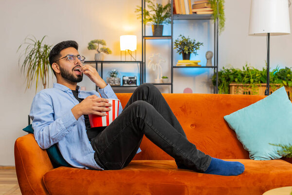 Relaxed Indian young man eating popcorn and watching movie wow expression lying on sofa in living room at home. Happy Hispanic guy in casual clothes enjoying film during weekend in apartment room.