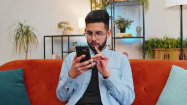 Akıllı telefonu elinde tutan heyecanlı Arap genç adam online bahis oyununun kazanmasıyla hayrete düşmüş iyi haberler okuyor. Kanepede oturan mutlu Hintli adam evdeki galibiyetten çok mutlu bir şekilde ekrana bakıyor..