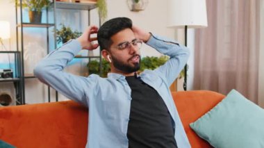 Kablosuz kulaklıklarıyla koltukta dinlenen mutlu, rahat Hintli genç adam çevrimiçi müzik listesinden ortamı yatıştıran sakin müzikler dinliyor. Gülümseyen İspanyol adam ellerini başının arkasına koyup odada dinleniyor..