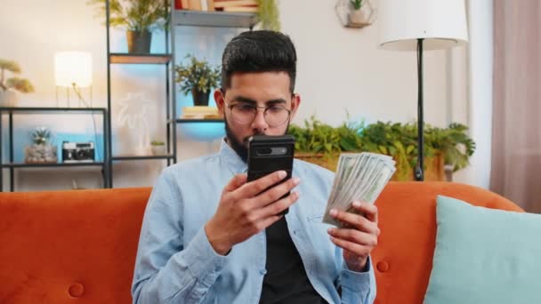 成功した金持ちのインド人男性がスマートフォンを持ち 家でソファーに座っているドル紙幣ファンに手を振った ハッピーアラビアの男は 予算の概念を計算するオンライン宝くじゲームの計画休暇に勝ちました — ストック動画
