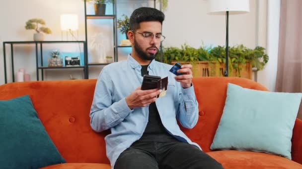 インターネットストアでオンラインショッピングで支払いを行う幸せな若いインド人男性 自宅でソファーでモバイルアプリで検証プロセスに関与する銀行クレジットカードを持っているヒスパニックの男笑い — ストック動画