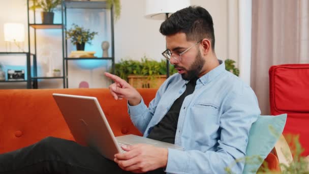 自宅でソファーに座っている若いインド人男性は 不安とストレスを持つラップトップを使用しています 衝撃を受けたアラビア人の男は リビングルームのアパートでネットブックで悪いニュースを失うことに反応しました コンピュータウイルス — ストック動画
