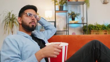 Rahat Hintli genç adam patlamış mısır yiyor ve evdeki oturma odasında kanepede oturup film seyrediyor. Hafta sonu dairede film seyreden günlük kıyafetli mutlu İspanyol adam..