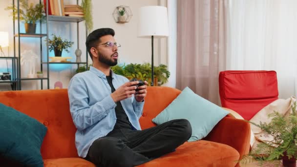 リビングルームでソファーに座って楽しむジョイスティックコントローラーを使用して陽気なインドの若者 自宅で週末にソファーでオンラインゲームで成功を収めているアラビアの男 — ストック動画