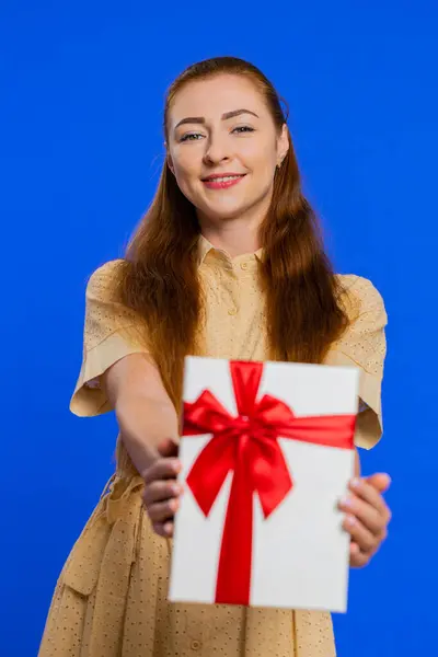 Positive smiling adult woman presenting birthday gift box stretches out hands, offer wrapped present career bonus, celebrating party. Redhead girl isolated on blue studio background indoors. Vertical