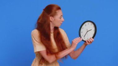 Endişeli genç bir yetişkin kadın, saati kontrol ediyor, işe geç kalıyor, gecikiyor, son teslim tarihi. Kızıl saçlı kız, mavi stüdyo arka planında dakikalarca yalnız kalmaktan endişeleniyor.