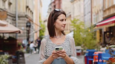 Akıllı telefon kullanarak gülümseyen genç bir kadın mesajları cevaplıyor internetteki görüntülü sosyal medya uygulaması ile sohbet ediyor. Kız turist ellerinde telefonla şehir sokaklarında yürüyor.