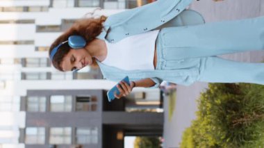 Kablosuz kulaklıklı, neşeli, çocukluk çağındaki mutlu çocuk dışarıda akıllı telefon dansında en sevilen disko rock n roll müziğini dinliyor. Şehir caddesinde yürüyen bir kız. Dikey