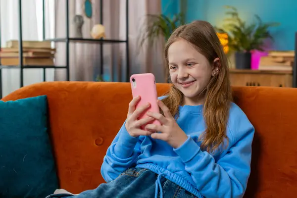 stock image Cheerful preteen girl texting share messages on smartphone social media applications online watching relax movie. Female Caucasian teenager kid using mobile phone at home in living room sits on sofa.