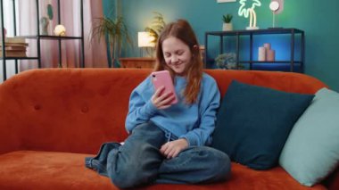 Neşeli genç kız, internet üzerinden akıllı telefon sosyal medya uygulamalarında rahatlama filmi izlerken mesajlarını paylaşıyor. Oturma odasında cep telefonu kullanan beyaz bir kız çocuk kanepede oturuyor..