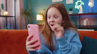 Akıllı telefon daktiloyla yazı yazan mutlu bir kız büyük bir sürpriz buldu, iyi haberler, online maç kazancını kutladı, yüzüne esen rüzgarı. Evdeki kanepede oturan genç bir kız.