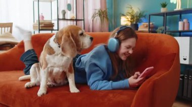 Kablosuz kulaklıklı mutlu beyaz kız çocuğu evde koltuğa uzanmış en sevilen enerjik disko dans müziğini dinliyor. Kanepenin yanında akıllı telefon kullanan gülümseyen çocuk..