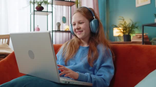 音楽やレッスン 遠隔学習にノートパソコンのリストを使用してヘッドフォンで19歳の少女 オンライン教育 笑顔の幼い子供がリラックスして ソファーに座っている家庭の職場で休憩を取る — ストック動画