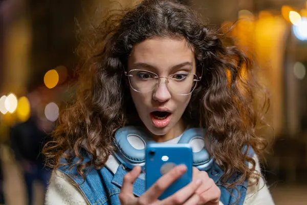 Oh my God Wow. Excited happy amazed excited winner young woman using smartphone typing browsing celebrating win good message news. Girl with brown windswept hair on urban city street at night evening