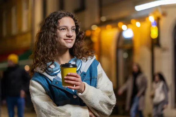 ハッピーブルネットの若い女性は コーヒーホットドリンクを楽しんで 遠く離れて考えている笑顔 リラックスして休憩 都会の街角のティーンエイジャーの少女は 屋外で夜にコーヒーを飲んでいる ライフスタイル ストック写真