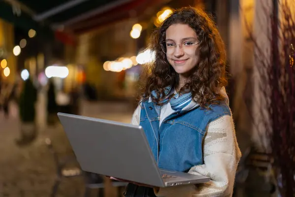 ハッピーティーンエイジャー少女は 街路のラップトップでオンライン遠隔の仕事をしています 屋外でチャットウェブサイトを閲覧 夜に町でタイピングを考えるカメラを見ている白人若い女性観光客 ストック画像