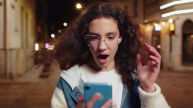 Aman Tanrım. Vay canına. Heyecanlı mutlu, neşeli genç bayan akıllı telefon daktilo kullanarak kutlama yapıyor ve iyi haber kazanıyor. Gece vakti şehir caddesinde kahverengi rüzgarlı saçlı bir kız..