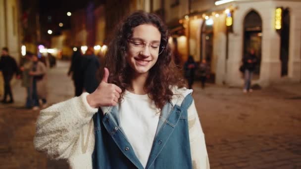 同じように ハッピーティーンエイジャーの子供は 肯定的な何か良いフィードバックをサインのように2つの親指を示すカメラで承認して見ています 夜の街路にいる少女 街のライフスタイル — ストック動画