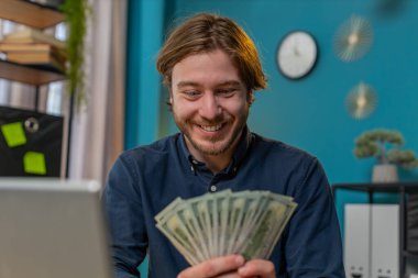 Harika bir iş adamı elinde nakit para banknotlarının hayranı. Başparmaklarını dizüstü bilgisayarına gösteriyor ve işaret ediyor. Kutlama, başarı, internet gelirleri, merkez ofisteki zenginlik. Uzak çalışma.