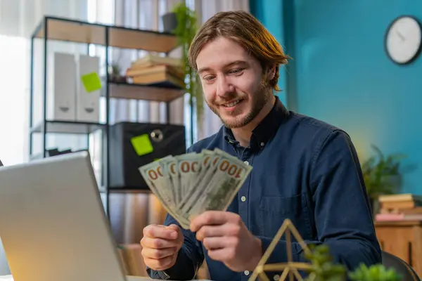 Happy businessman guy holding fan of cash money dollar banknotes showing thumbs up and pointing at laptop, celebrate, success career, online income, wealth at home office desk. Remote distant working.
