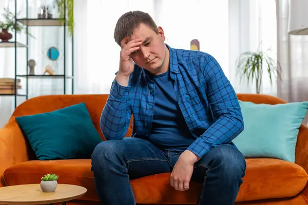 Sad lonely Caucasian man headache looks pensive thinks over life concerns suffers from unfair situation sits on sofa. Upset guy with hand on head depressed feeling bad annoyed in living room at home.