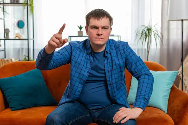 Quarrel. Displeased aggressive unhappy Caucasian young man gesturing hands with irritation and displeasure, blaming scolding for failure, asking why this happened. Portrait of guy sitting on home sofa