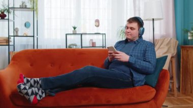 Kablosuz kulaklıklı mutlu beyaz adam akıllı telefondan en sevilen enerjik müziği dinliyor. Hafta sonları evdeki oturma odasındaki kanepede uzanmış gülümseyen erkek..