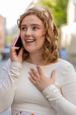 Mutlu, gülümseyen, kızıl saçlı, uzaktan iletişim kuran genç bir kadın dışarıda akıllı telefondan konuşuyor. Şehir caddesinde yürüyen bir kız beklenmedik bir şekilde iyi haber dedikodusu yapıyor. Kasaba yaşam tarzları