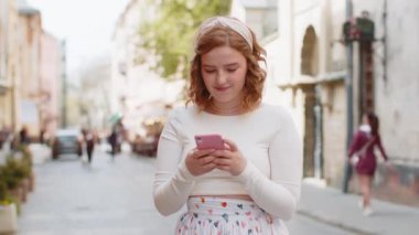 Uzun elbiseli, akıllı telefon kullanan, sosyal medya mesajları yazan açık hava gezici uygulamasında haritada bir yol arayan kızıl saçlı genç bir kadın. Şehir caddesinde yürüyen bir kız. Kasaba yaşam tarzları