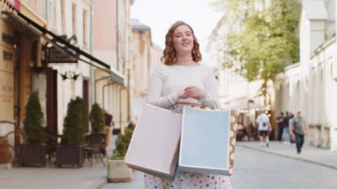 Alışveriş merkezinde açık havada hediyelerle dolu poşetlerle alışveriş yaptıktan sonra uzun elbiseli, mutlu, kızıl saçlı genç bir kadın. Şehir caddesinde yürüyen turist kız. Kasaba yaşam tarzları.
