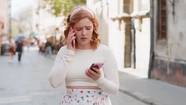 イライラした神経症は 2つの携帯電話で叫ぶ赤毛の若い女性が会話の紛争の争いの論争を屋外で解決する問題を議論することを強調しました 都市のストリートの少女 — ストック動画