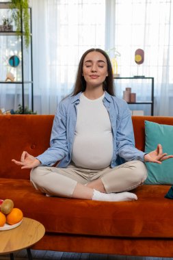 Evde, oturma odasında rahat bir kanepede meditasyon yaparken nilüfer pozisyonunda oturan hamile, çekici bir kadın. Sağlık, hamilelik ve nefes alma pratiği kavramı. Mutlu bayan anne olmaya hazırlanıyor.