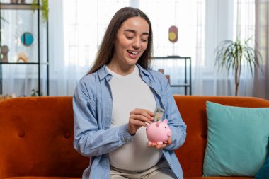 Mutlu, genç, hamile bir kadın kumbaraya banknot sokuyor ve evdeki oturma odasındaki kanepede oturarak koca göbeğini okşuyor. Kanepede bebeklerin gelecekteki konsepti için para biriktirirken gülümseyen anne kadın.