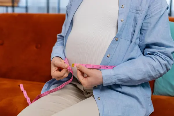 Unrecognizable pregnant woman sits on sofa measuring belly diameter with measure tape. Future mother expecting awaiting childbirth monitoring body changes weight gain dimensions. Pregnancy maternity