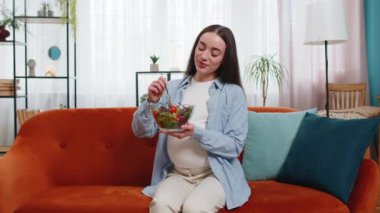 Mutlu, iştahlı hamile bir kadın oturma odasının iç tarafındaki kanepede oturmuş sebze salatası yiyor. Annelik kendi kendine bakım. Gülümseyen beyaz kız doğru sağlıklı vejetaryen yemeğini seçiyor.