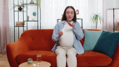 Evdeki oturma odasındaki kanepede oturan, doğmamış çocuğu beslemek için uyuşturucu ve içme suyu alan hamile bir beyaz kadın. Sağlık sigortası ve hamilelik kavramı. Bayan mutlu bir şekilde kameraya bakıyor..