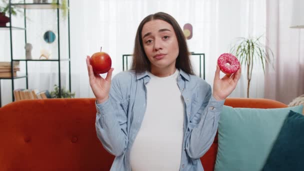 选择正确的营养 困惑的年轻孕妇比较甜甜的甜甜圈和成熟的苹果 耸耸肩在不确定性中 未来的母亲不愿在甜点和水果之间做出选择 — 图库视频影像