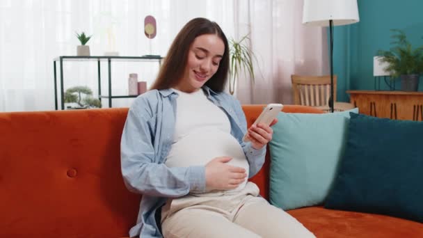 在家里客厅里 坐在沙发上聊天 用智能手机发短信给社交媒体发笑的孕妇 快乐的白种人女士未来的妈妈抱着手机在公寓里抚摸着肚子 — 图库视频影像