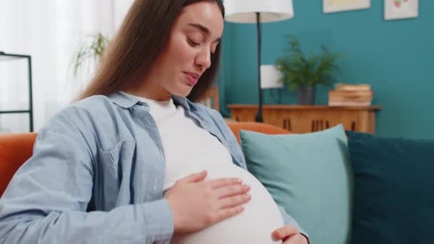 妊娠中の女性はラビーを歌い 自宅のリビングルームでソファーに座って大きな腹に触れました 将来の母親の女性を愛し ソファーで余暇を過ごす未生児の赤ちゃんと話しています 新しいファミリーコンセプト — ストック動画