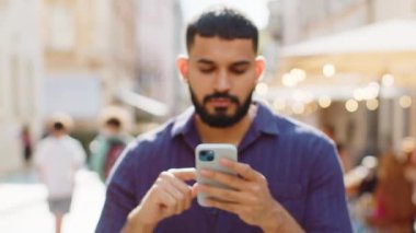 Akıllı telefon kullanarak gülümseyen Hintli adam internete mesajlar yazıyor, işi bitiriyor, dışarıdaki kameralara bakıyor. Şehir caddesinde yürüyen bir adam. Kamera yakınlaşıyor ve kişi odaklanıyor