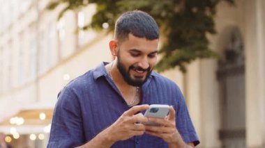 Hintli genç adam cep telefonuyla kutlama yapıyor iyi haber kazan, piyango zaferi kazan, açık havada dağıtım yap. Şehir caddesinde yürüyen mutlu yetişkin erkek turist. Kasaba yaşam tarzları