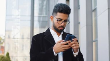 Mutlu Hintli işadamı akıllı telefon yazıcı kullanıyor e-posta mesajlarına cevap veriyor açık havada online mobil uygulamalarla sohbet ediyor. Şehir merkezindeki kameraya bakan Arap Hintli adam. İş adamları
